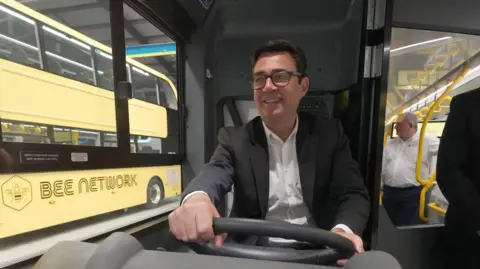 PA Greater Manchester mayor Andy Burnham smiles as he drives a Bee bus - a yellow double decker bee bus is out of the window