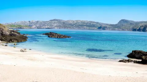 A sandy beach on Handa on a beautiful sunny day. The shallow sea water reflects the blue of the sky above. On the opposite shore are low rocky hills.
