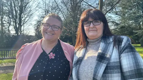 North Yorkshire Council Two women, both with dark hair and glasses, stand side by side in the sunshine. One is wearing a pink cardigan with a flower necklace and a black top with light spots. The other is wearing black-and-white shirt on top of a beige jumper.    