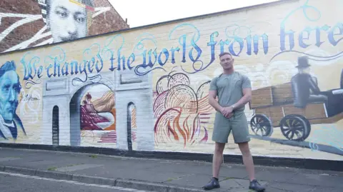Luke Jenk is wearing shorts and a t-shirt and is standing in front of a mural on a wall in Darlington. It reads We changed the world from here and it celebrates the railway history of the town.
