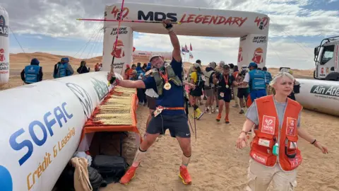 A picture of Marc Lockwood at the finish of the Marathon des Sables across the Sahara Desert. Marc is smiling and holding up two walking sticks in a cross shape.