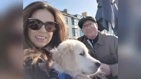 Roma Morgan Roma and Steve Morgan photographed with Kobi. Roma is holding Kobi who is a puppy at the time the picture was taken. She is wearing sunglasses, has long brown hair, and is smiling at the camera. Steve is stood next to them. He is wearing glasses and a flat cap.
