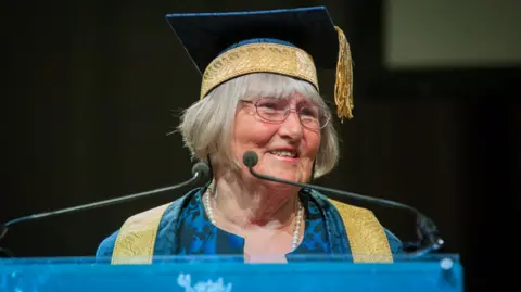 An older lady with a grey bob wearing a floral teal mortarboard and gown with a gold tassel smiles as she speaks into microphones at a university graduation ceremony. She is also wearing thin-framed silver glasses and a pearl necklace.
