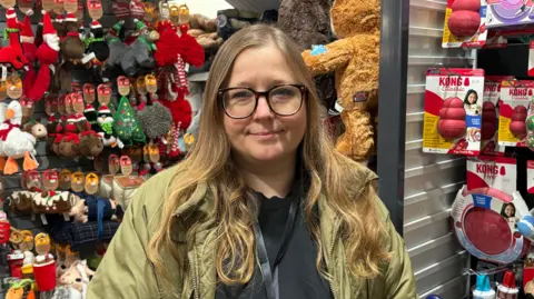 BBC Anna has long light brown hair and wears a light green jacket and black top. She stands in front of a display of Christmas related toys in a shop. 