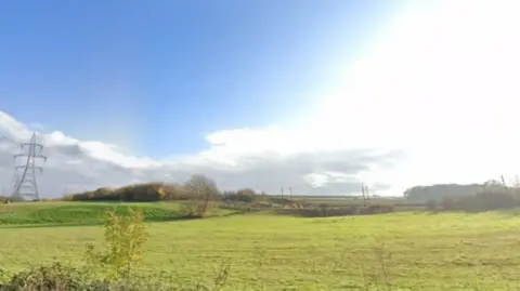 A green field with energy pylons in the left of the image. The field is vast with trees in the background.