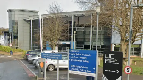 A view of the campus of the University for the Creative Arts in New Dover Road, Canterbury, featuring many steel and concrete buildings, a sign announcing the name of the institution and a blue and white sign giving directions to different parts of the campus.
