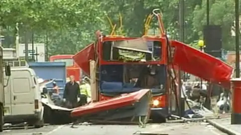 A red London bus with metal and debris protruding from what was once the roof, with emergency workers nearby.