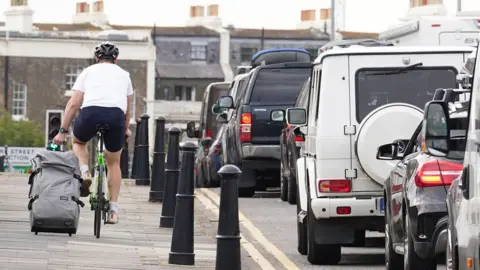PA Media A man wearing a white T-shirt and a helmet cycles past a long line of traffic.