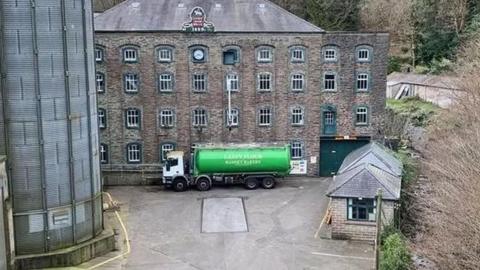 A small office block set in front of a mill building with a green flour truck parked in front of it and a flour silo to the left hand side