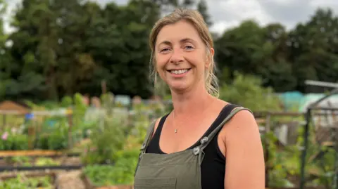 Janet Wood smiling at the camera, is in a black strappy top with a green gardening apron over the top.