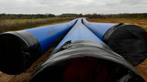 A view of three large blue water pipes that span off into the distance. They sit in a field and their openings have been covered by black sheeting. Grass and fields can be seen either side of the pipes.