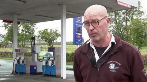 Ian Chambers. He is wearing a maroon jumber containing the logo for Chambers Garage. He is bald and is wearing glasses. He is standing in front of his petrol station.