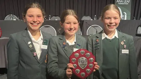 BBC Three young girls in green blazers smiling and Sienna holding a plaque which has silver on it.