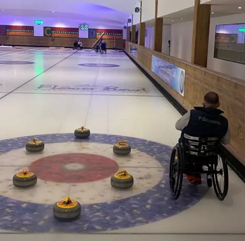 LDRS A session taking place on the ice rink in Barton. The photo is taken from the back of the rings - bue white and a red bullseye with six yellow curling stones in a circle on it. To the right Paul Simmons is in a wheelchair with his back to the camera and players can be seen in the distance about to play