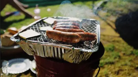Getty Images The picture shows a disposable barbeque placed on a bucket. It has a silver casing, and silver wire goes across the top in a criss-cross pattern. Sausages are cooking and there is smoke coming from them. There are various bowls, plates and cups in the background.