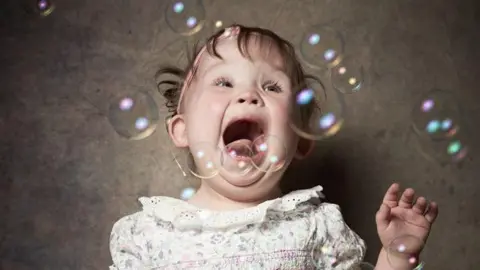 Elizabeth, a young girl with short dark hair, a pink headband and in a floral dress, screams with excitement as bubbles float around her head.