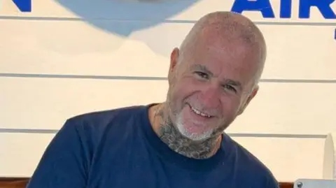 Family handout Simon Beston smiling at the camera. He has very short shaved grey hair and beard. He is wearing a blue t-shirt and has a neck tattoo. 