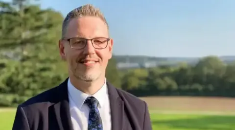 West Mercia Police and Crime Commissioner A man with short, spiked greying hair is wearing dark-framed glasses, a black suit, white shirt and dark patterned tie. He is standing in front of a blurred background of rolling fields and trees with a more urban landscape behind. 