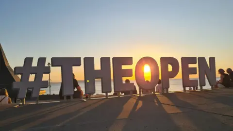 A huge sign for The Open with a hashtag beside it. The sun is behind the sign and there are people sitting around it.