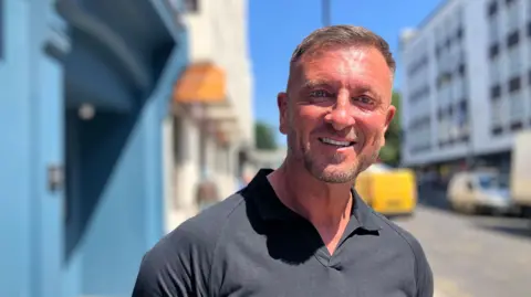 A man, Jason Burrill, stood on a pavement by a street.