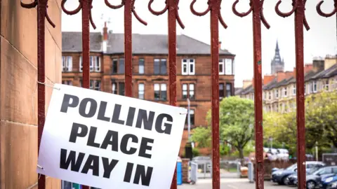 PA Media Sign reading “Polling place way in” attached to a red metal gate outside a brick building, with residential buildings and trees visible in the background.