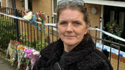 Robby West/BBC Rona Wood wears a black fluffy jacket and stands against the railing of a house. A police cordon runs along the railings and flowers have been left at the base of the railings. 