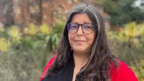 Simon Dedman/BBC A woman with long brown and grey hair is looking at the camera. She is wearing black frame glasses and a red cardigan and a black shirt.