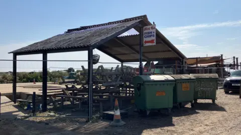 Guy Campbell/BBC Stacked up seating benches and green wheelie bins can be seen at the area which was destroyed by fire last May