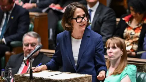 House of Commons Liz Kendall  has brown hair down to her shoulders and black-rimmed glasses. She is speaking at a podium in the House of Commons and is wearing a dark blue suit jacket and white top.  