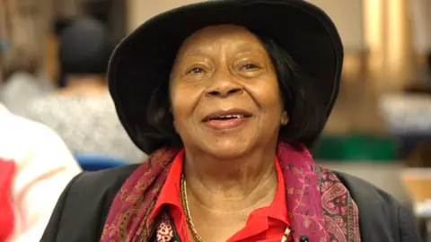 A black woman wears a large brimmed hat, red shirt and patterned red scarf