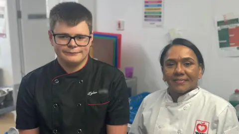 BBC Tyler, left, has short dark hair and is wearing dark rimmed glasses and a black chef's uniform. Anna has dark hair pulled back in a pony tail and is wearing a white chef's uniform.