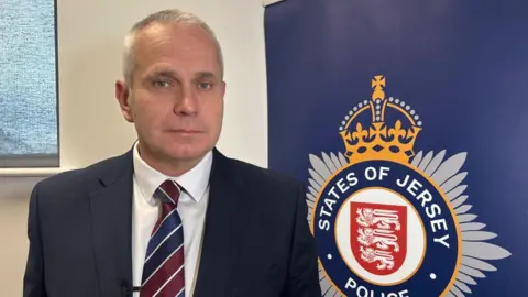 BBC A man in a blue suit and white top and blue and red tie. He is stood in front of a States of Jersey Police sign. The wall behind him is cream. 