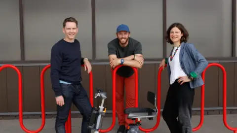 Supersmith Tom, Fred and Emily, all smiling into camera, standing by railings with their 3Scooter in the foreground