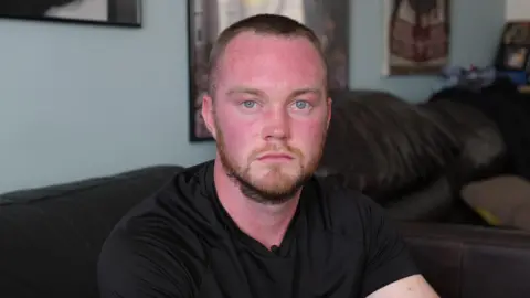 Geraint is facing the camera, wearing a black t-shirt sitting in his living room.