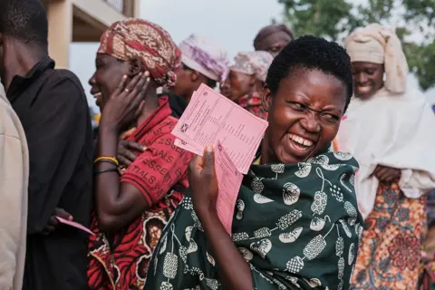 TCHANDROU NITANGA / AFP / GETTY IMAGES A woman jests as her photo is taken in the voting queue at a primary school in the capital, Gitega. She is holding several ballot papers in one hand. 