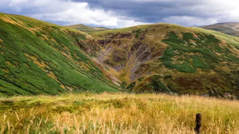Christina Cox A broad landscape of green and golden hills surrounds a steep-sided valley beneath dramatic, cloud-filled skies.