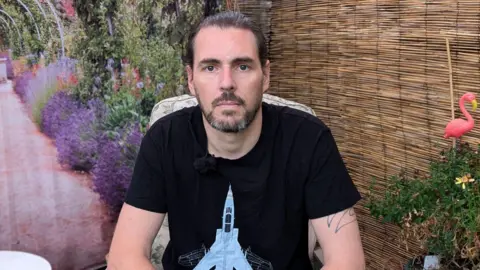 A bearded Lee Windell sitting outside in a garden surrounded by colourful plants and wearing a black t-shirt with an aeroplane design