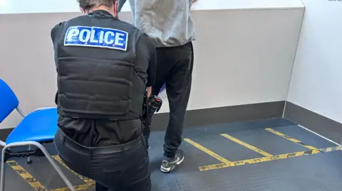BBC/ Kate Bradbrook Police officer with his back to the camera searches the socks of a man who has been brought into custody. Their faces cannot be seen.