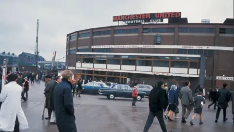 BBC Fans, wearing coats in light rain, walk towards Old Trafford during a match day in 1967