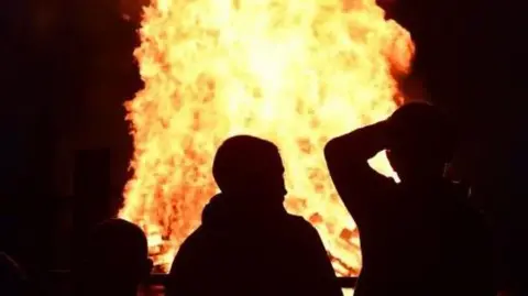 Three young people's silhouettes can be seen standing in front of a lit bonfire. 
One young person has their hand on the head and the other two are looking at them.
