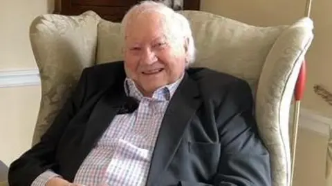 An older man with white hair sits in a high-backed armchair, smiling into the camera. He is wearing a checked shirt and a black blazer jacket.