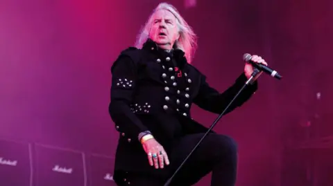 Getty Images A man with long blonde hair dressed in black on a stage with a microphone.