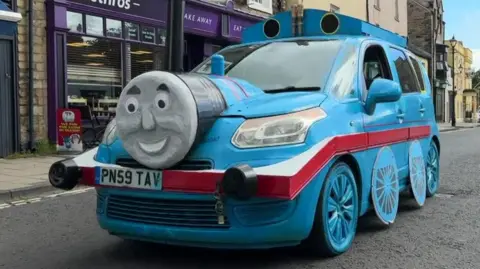 A wide shot of the bright blue four-door car as it drives through Haltwhistle complete with Thomas the Tank Engine frontage.