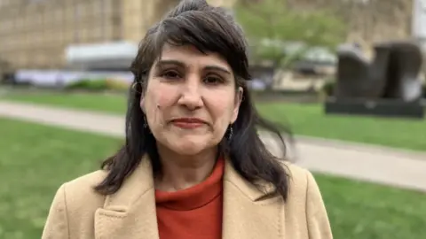 Dame Jasvinder Sanghera looking at the camera. She has dark, shoulder-length hair and is wearing red lipstick, a red top and a beige jacket. There is path and grass in the back ground which is blurred.