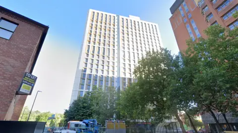 A city street lined with tall buildings, including a modern white‑and‑glass high‑rise, an older brick building with a large “To Let” office‑space sign, and a reddish‑brown tower. Trees partially obscure the view, with construction vehicles and equipment at street level.