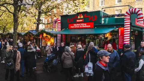 Teamjackson/iStock/Getty People walk past and queue at various stalls during York Christmas Markey, with attendees wrapping up in warm winter clothing. A mulled wine and hot chocolate stand is serving customers, with smaller craft stands nearby. 