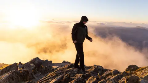 ConorHikes A man is standing on the rocky summit of a mountain wearing a coat. Behind him are cloud inversions lit up by sunrise.