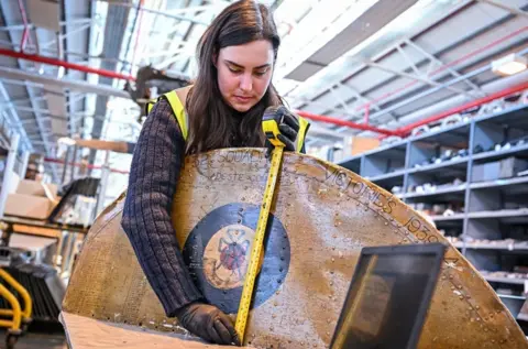 RAF Museum Someone measuring a large, mainly brown, object with a yellow tape measure. Shelves are in the background.