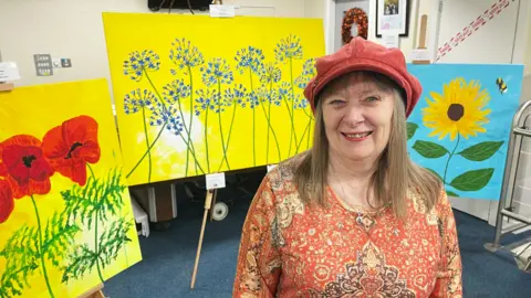 A woman in a red baker boy hat and a long sleved top covered in an Indian pattern is standing in a reception area. Behind her are three huge paintings on easels. They are all very bright. One is of three poppies on a yellow background. Another is of two sunflowers on a blue background and includes a bumblebee. The largest one is of agapanthus on a yellow background.