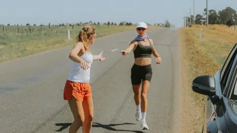 Megan Chapple Megan Chapple hands a Garmin device to another team member completing the run in Tasmania. The terrain is flat and it is a sunny day. A car is visible to the right.